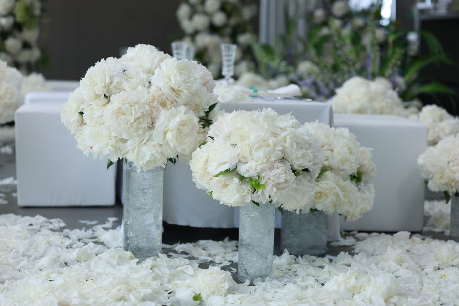 A floral arrangement featuring large, lush bouquets of predominantly white peonies and roses, arranged in tall, cylindrical glass vases with a textured surface. The flowers exhibit soft, full petals a