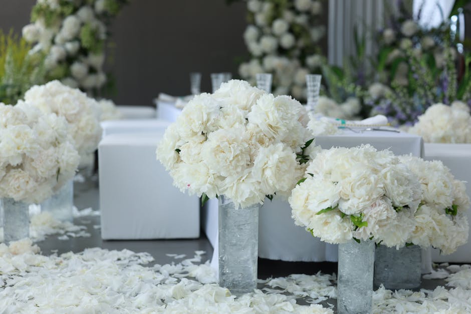 A floral arrangement featuring large, lush bouquets of white hydrangeas and roses, arranged in simple, cylindrical glass vases placed on a table covered with white petals. The flowers are fresh, with