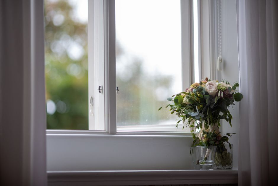 A beautifully arranged floral bouquet placed in a clear glass vase on a white window sill inside a room. The bouquet features a mix of soft pink and creamy white roses, complemented by green eucalyptu