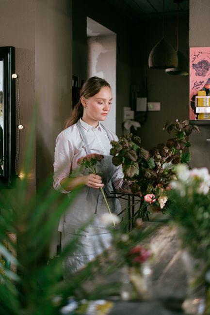 A young female florist with fair skin and light brown hair tied back, wearing a white blouse and an apron, arranging a bouquet of dark purple and pink flowers in a well-lit floral shop. She is standin