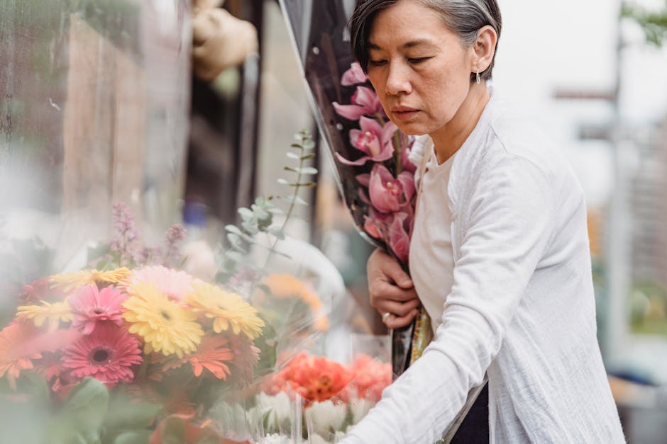 A woman with short dark hair, wearing a light grey cardigan over a white top, is holding a bouquet of pink orchids while selecting or arranging a floral display at a flower shop. The display behind he